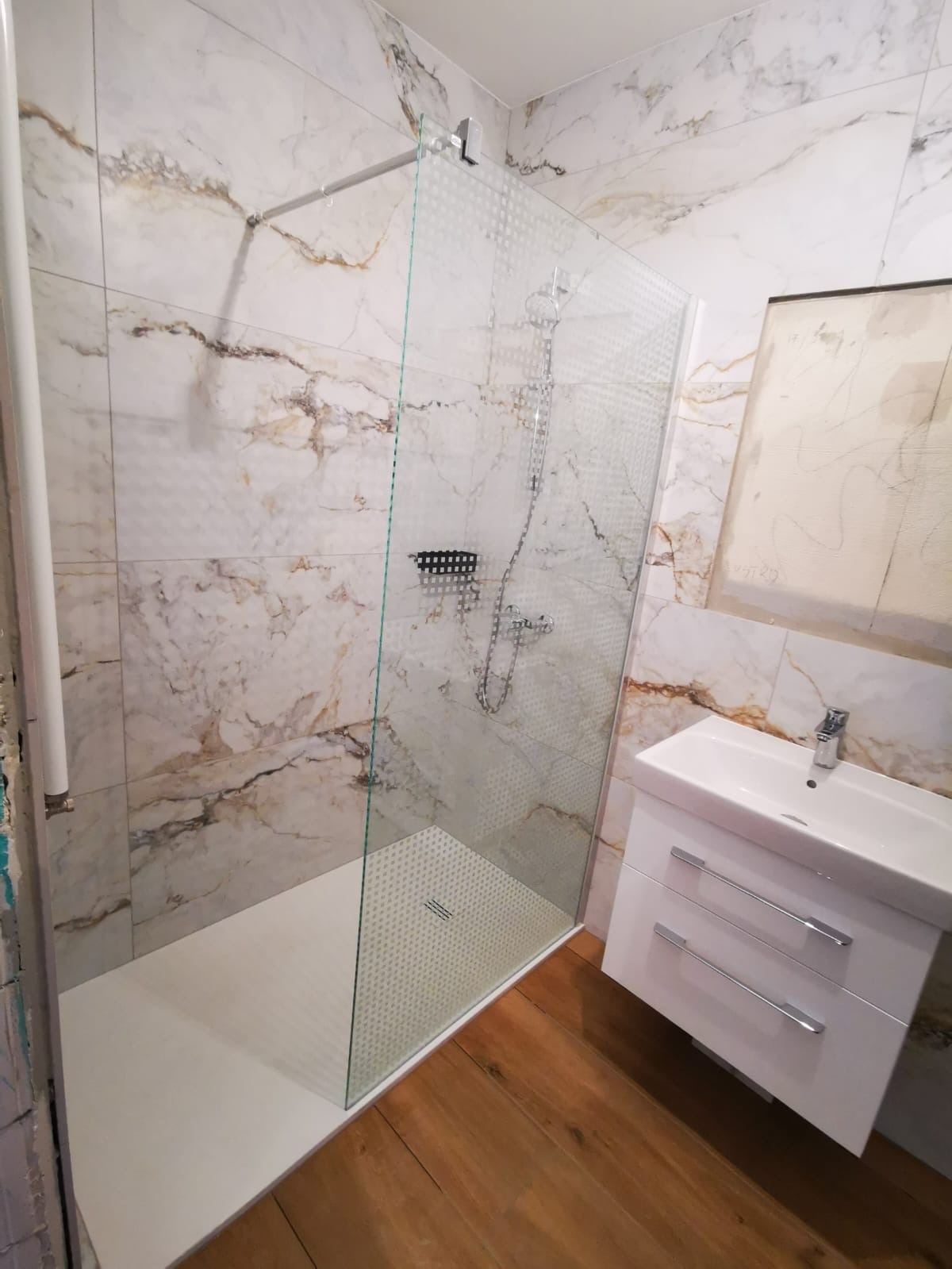 Wykończona łazienka w Oleśnicy: Kabina prysznicowa z transparentną ścianką z delikatnym wzorem, marmurowe płytki ścienne w odcieniach bieli i beżu, biała szafka pod umywalką z chromowanymi...