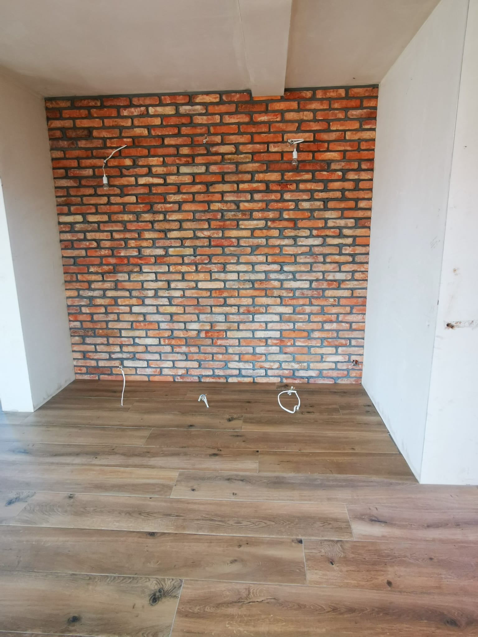 Ściana wyłożona dekoracyjną cegłą w trakcie prac wykończeniowych, widoczne wystające przewody elektryczne i świeżo położona podłoga imitująca drewno.