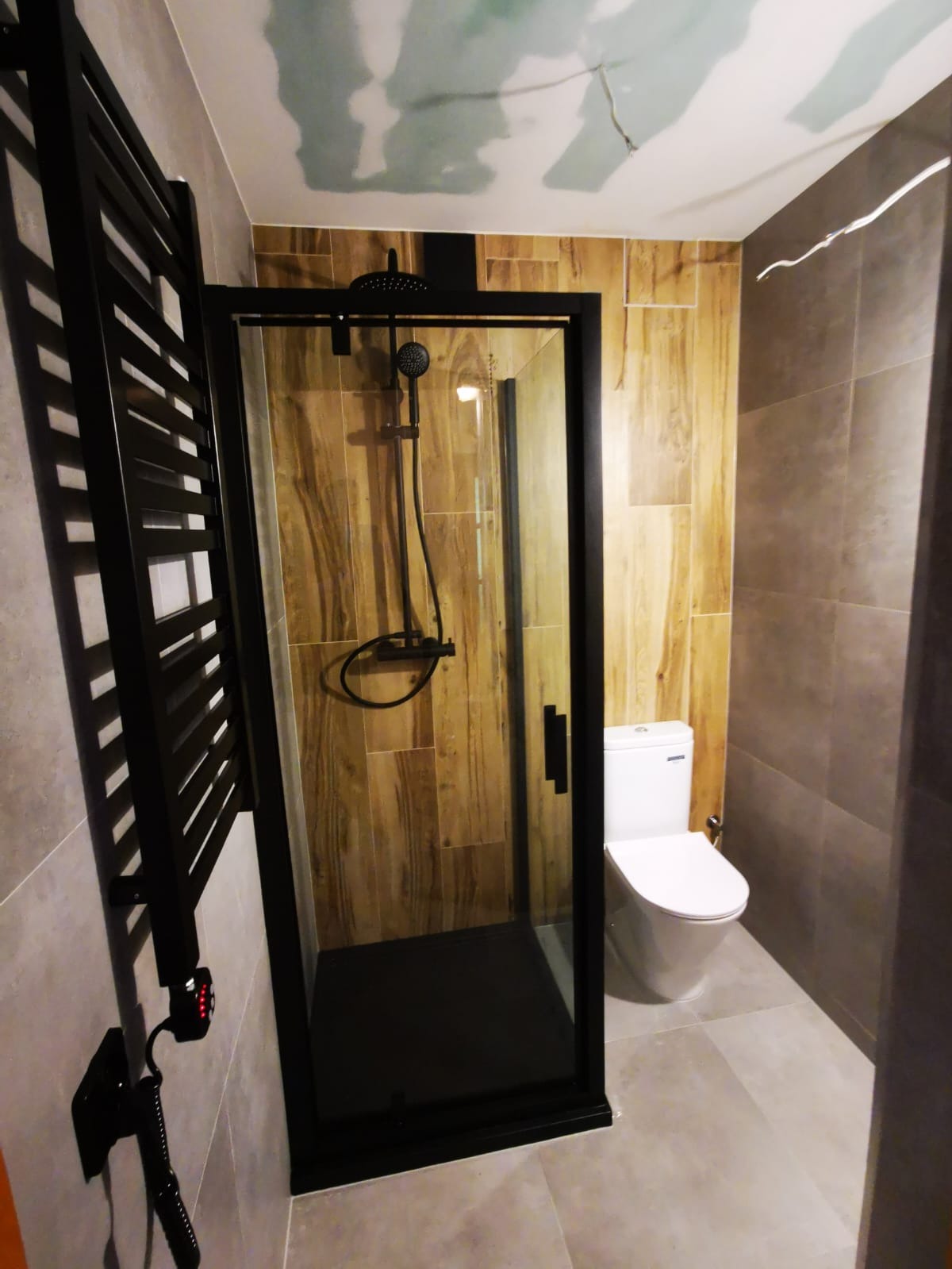 Nowoczesna łazienka z kabiną prysznicową w czarnej ramie, drewnopodobnymi płytkami na ścianie prysznica i szarą ceramiką na pozostałych ścianach, widoczny grzejnik drabinkowy i toaleta.