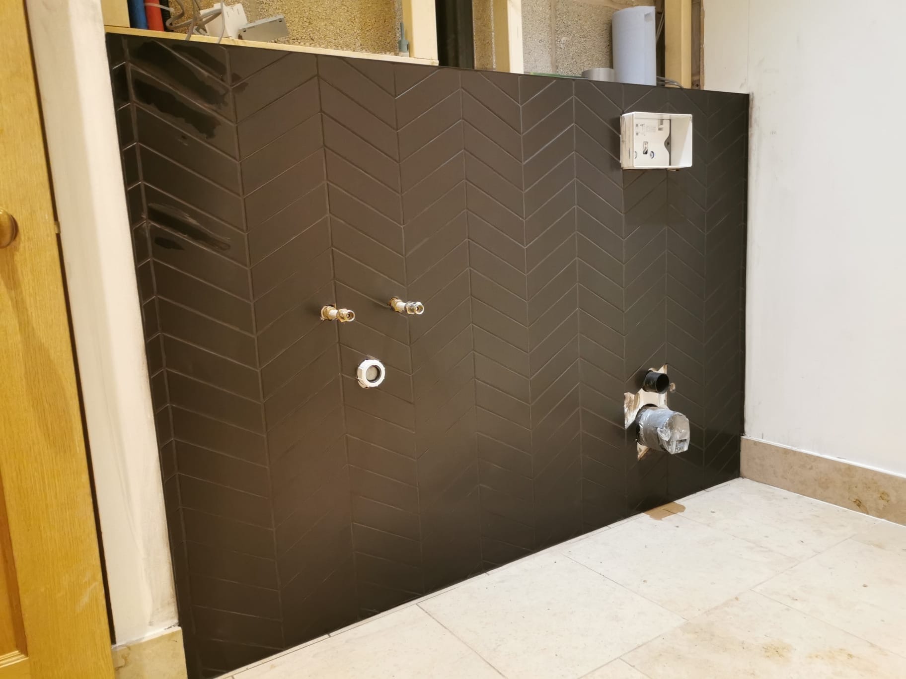 Ciemnobrązowe płytki ułożone w jodełkę na ścianie łazienki w trakcie remontu; widoczne przygotowane przyłącza wodne i kanalizacyjne, oraz puszka elektryczna podtynkową.