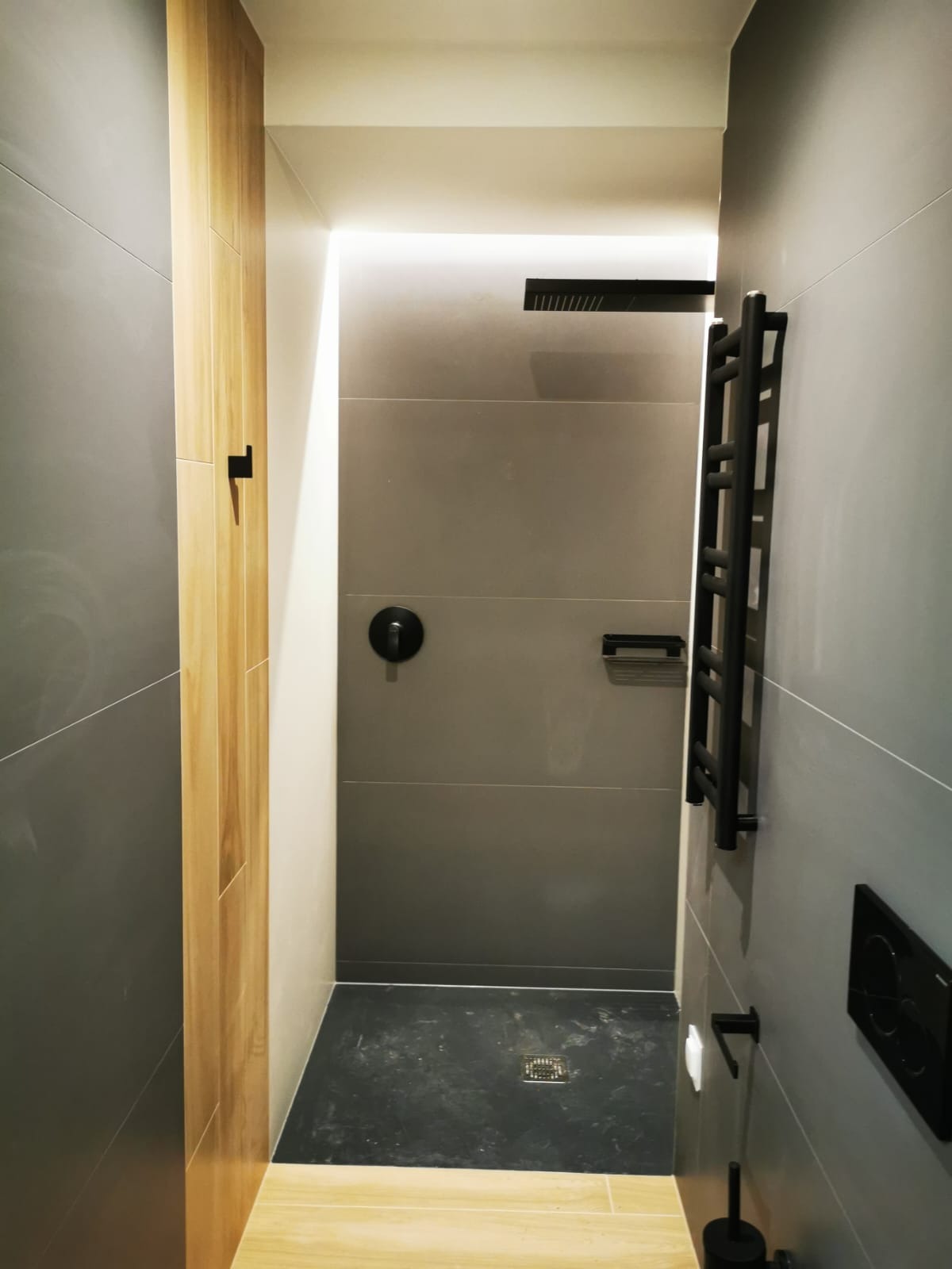 Nowoczesna łazienka z szarymi płytkami, czarną armaturą i drewnianym akcentem, oświetleniem LED w suficie i grzejnikiem drabinkowym.