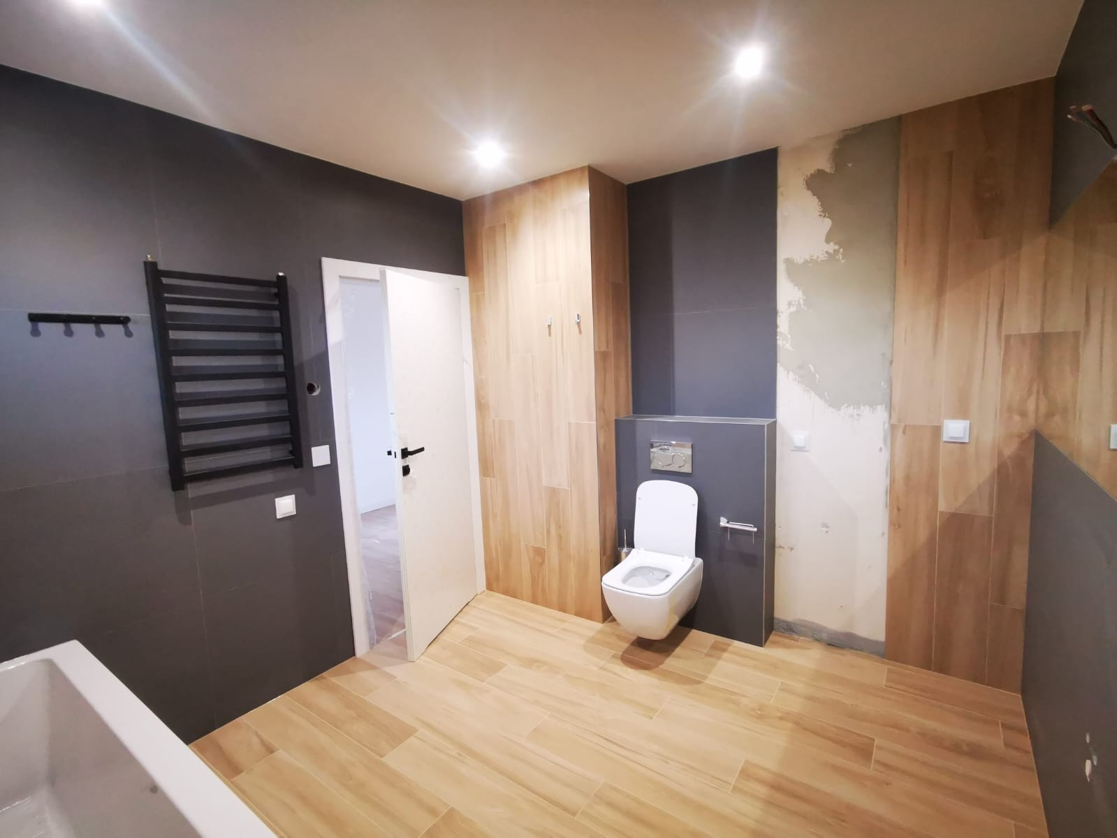 Wnętrze łazienki w trakcie prac wykończeniowych: drewnopodobne płytki na podłodze i części ścian, szara ściana z grzejnikiem drabinkowym, biała toaleta podtynkowa, fragment wanny, ściana w trakcie...