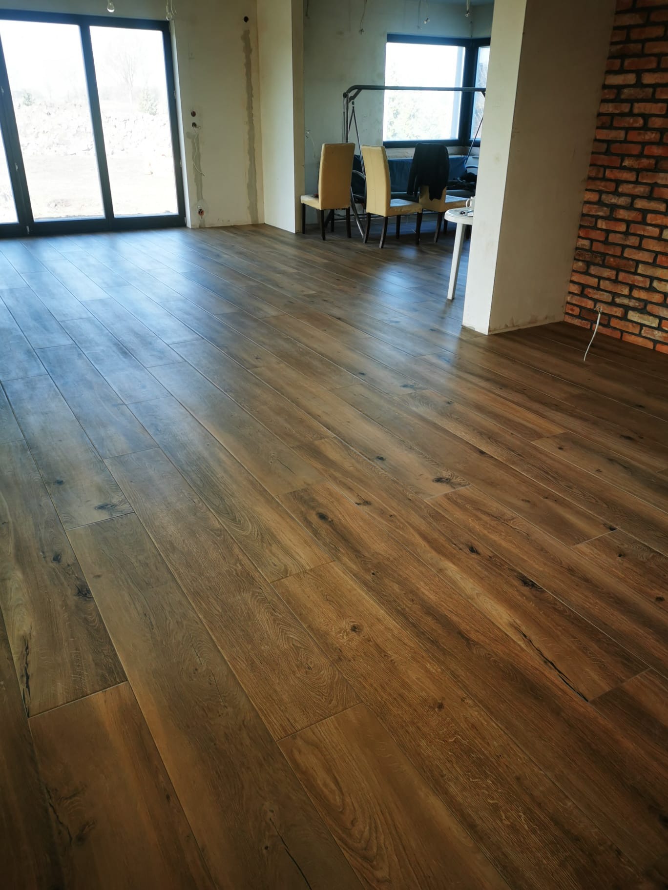 Parkiet ułożony w przestronnym, surowym wnętrzu z ceglaną ścianą i dużymi oknami, widoczne krzesła w tle.