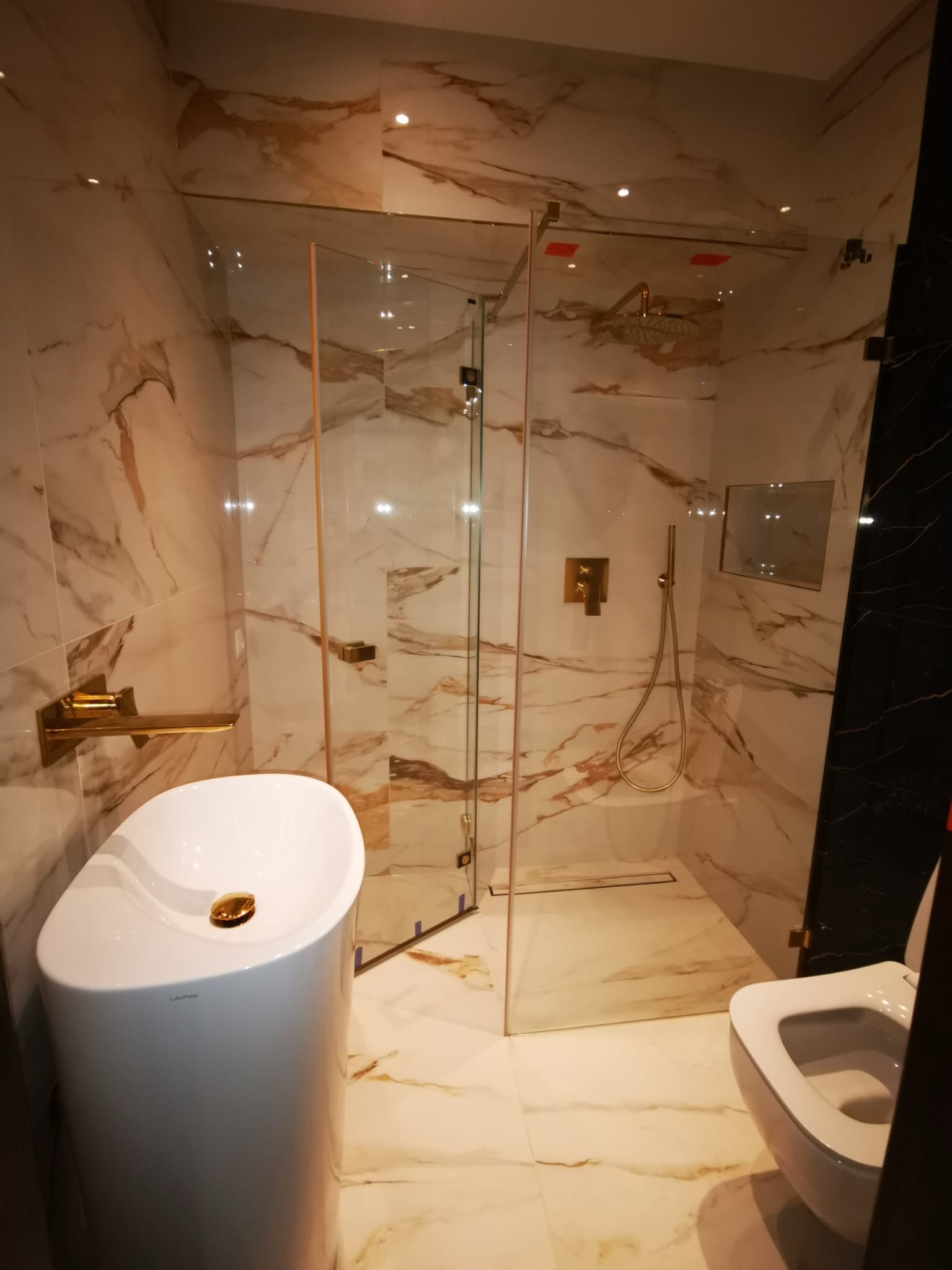 Nowoczesna łazienka z marmurowymi płytkami, kabiną prysznicową typu walk-in z złotymi detalami, wolnostojącą białą umywalką z złotym korkiem i toaletą.