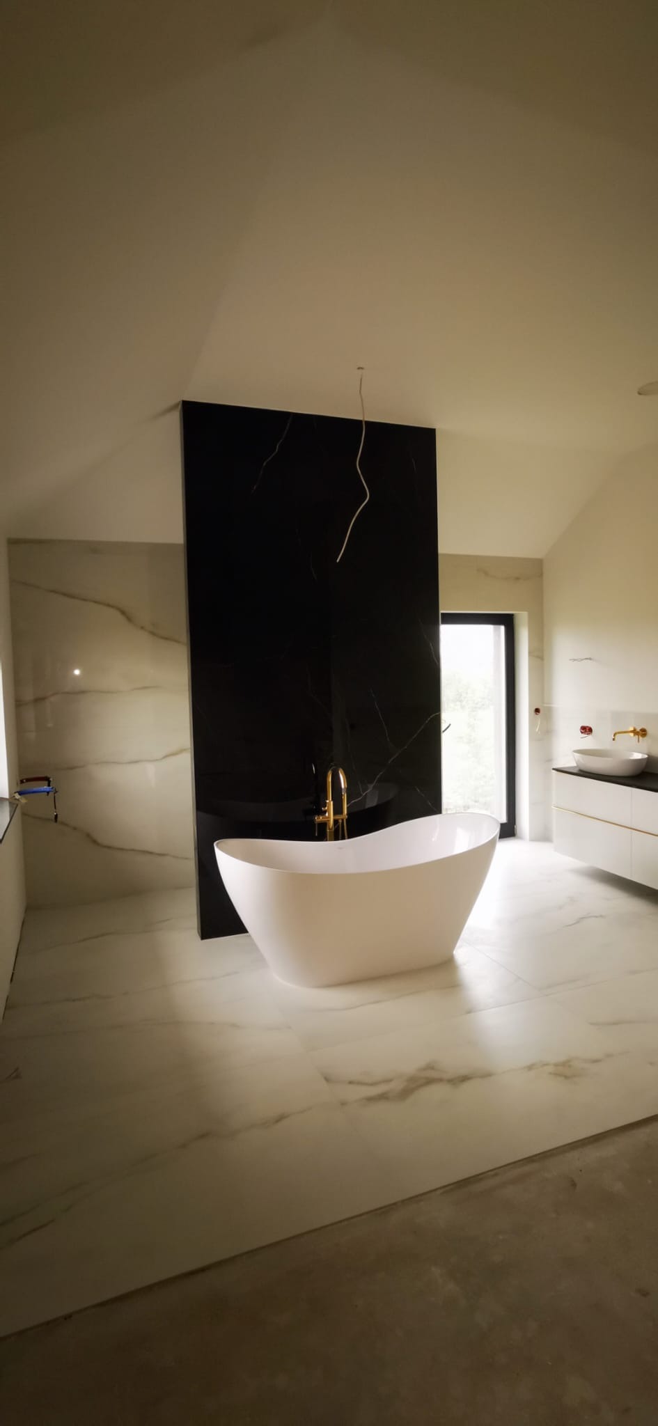 Nowoczesna łazienka z wolnostojącą białą wanną i złotą armaturą na tle czarnej płyty imitującej marmur, białe płytki podłogowe z delikatnym wzorem.