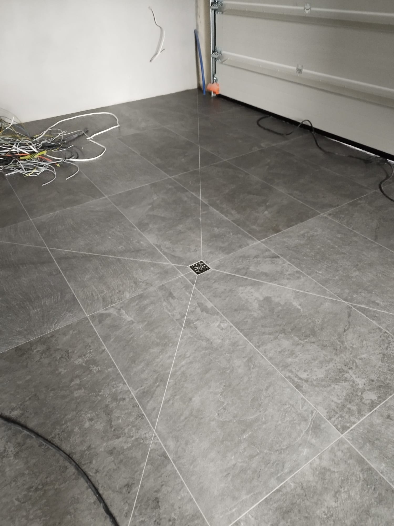 Wykończona podłoga w łazience z szarych płytek ceramicznych ułożonych w kwadraty, z centralnym odpływem liniowym, widoczne kable i przewody elektryczne na podłodze i ścianie, oraz brama garażowa...