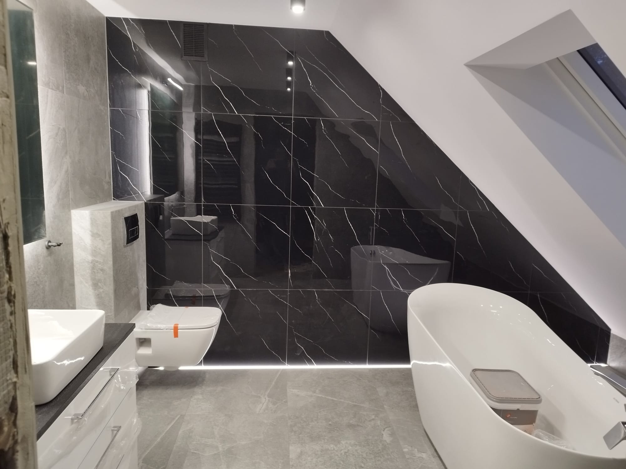 Nowoczesna łazienka na poddaszu z czarnymi płytkami imitującymi marmur na ścianach, wolnostojąca biała wanna, podwieszana toaleta z pomarańczową taśmą zabezpieczającą oraz biała umywalka z szafką.