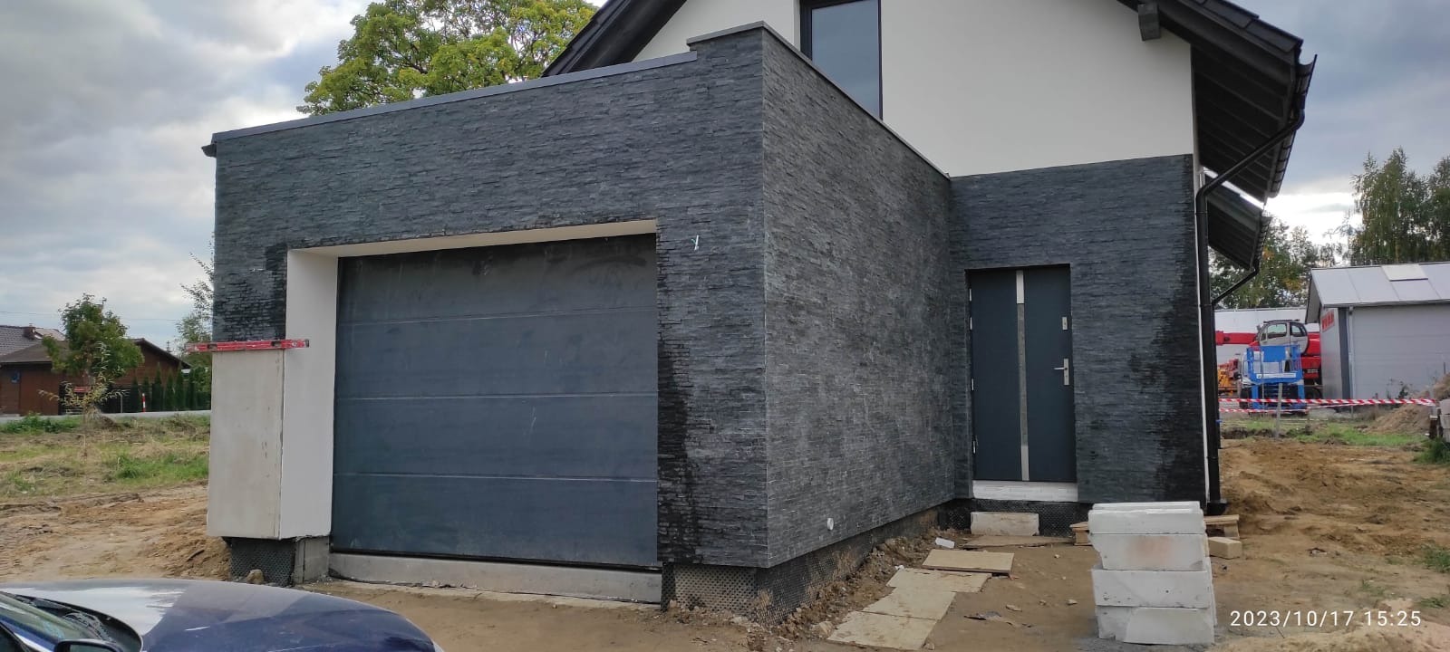 Elewacja budynku wykończona czarnym kamieniem łupkowym, widoczna brama garażowa i drzwi wejściowe, teren wokół w trakcie prac wykończeniowych.
