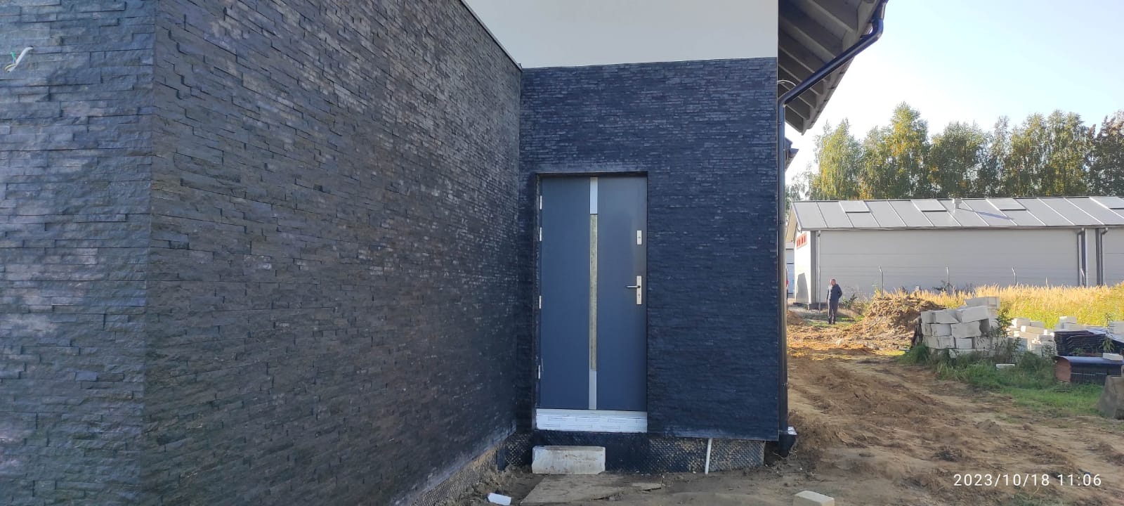 Elewacja budynku wykończona czarnym kamieniem łupkowym, z widocznymi drzwiami wejściowymi o stalowej ramie, na tle niezagospodarowanego terenu z budynkiem gospodarczym w tle.