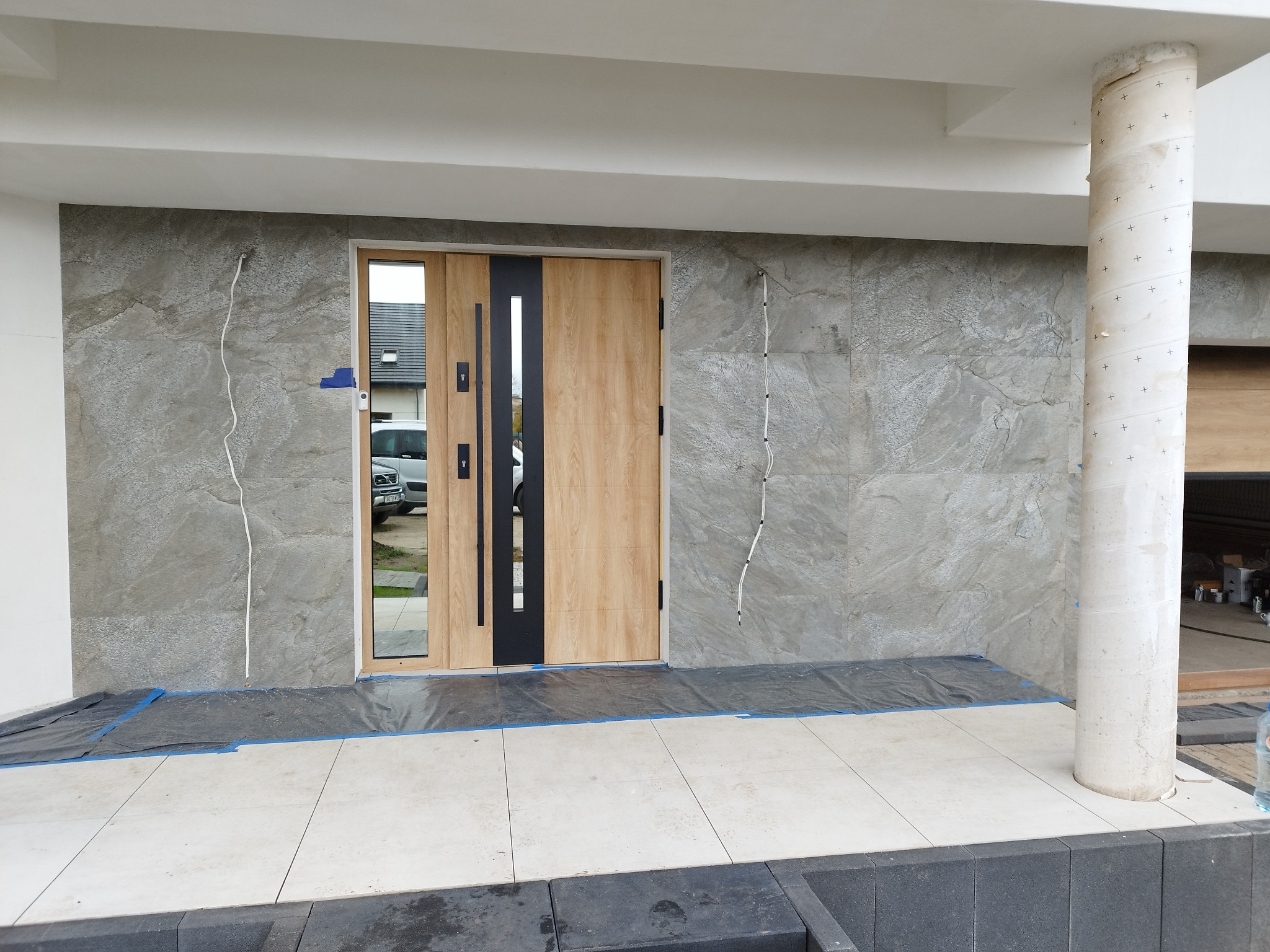 Montaż okładziny z płytek imitujących kamień na elewacji domu, widoczne kable i zabezpieczona podłoga przed wejściem.