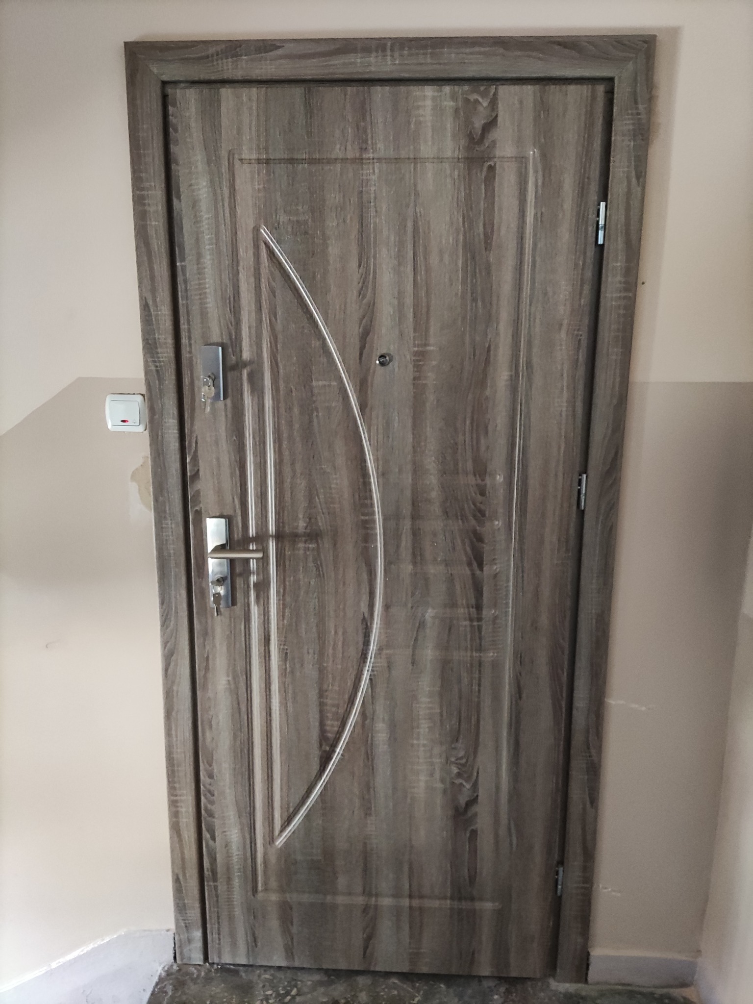 Zamknięte, nowoczesne drzwi wejściowe w odcieniach szarości z metalową klamką i wkładką zabezpieczającą, widoczne klucze w zamku, na ścianie obok włącznik światła.