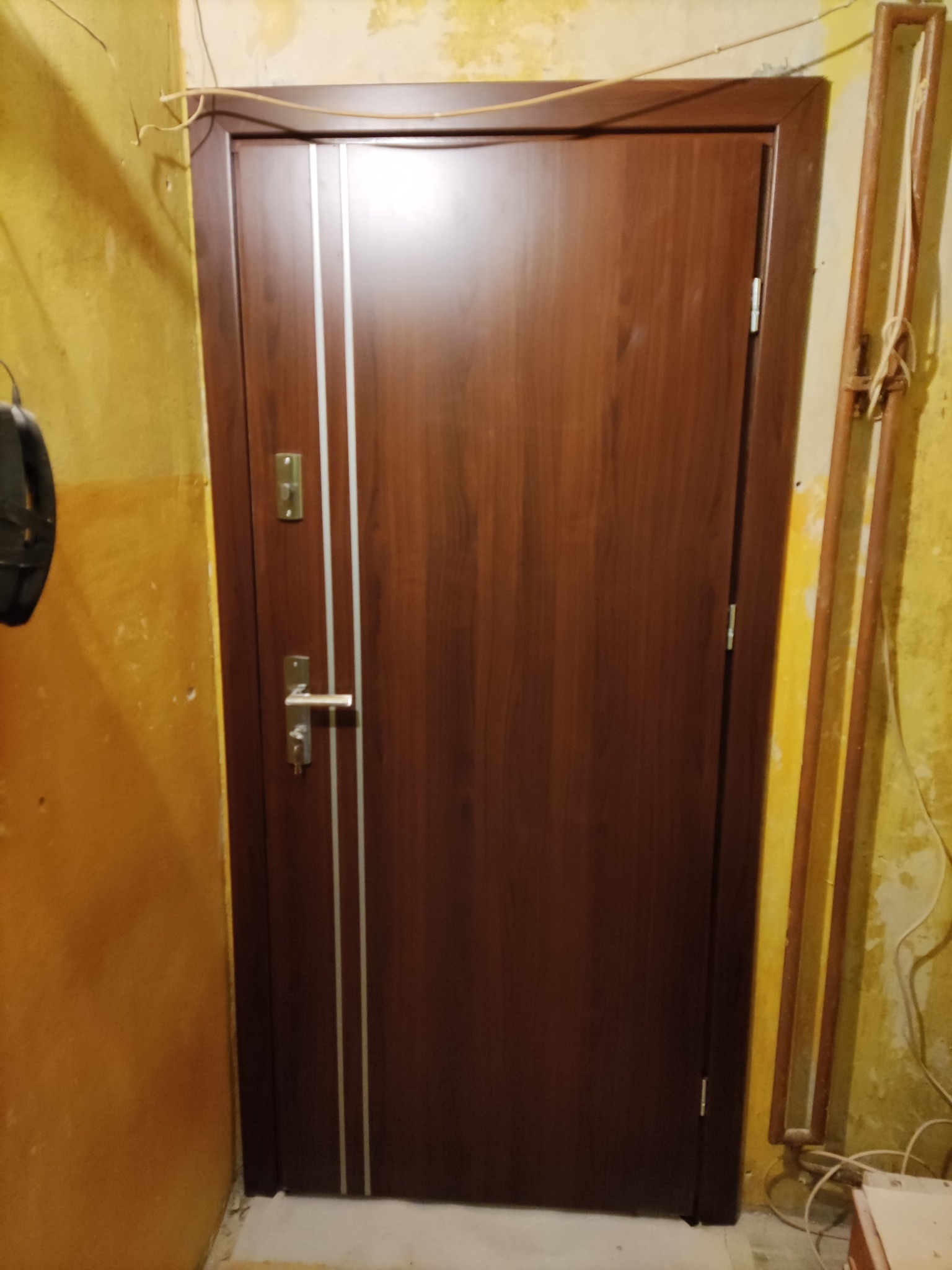 Ciemnobrązowe drzwi wejściowe z dwoma srebrnymi paskami pionowymi, zamontowane w korytarzu z żółtymi ścianami i widocznymi instalacjami.