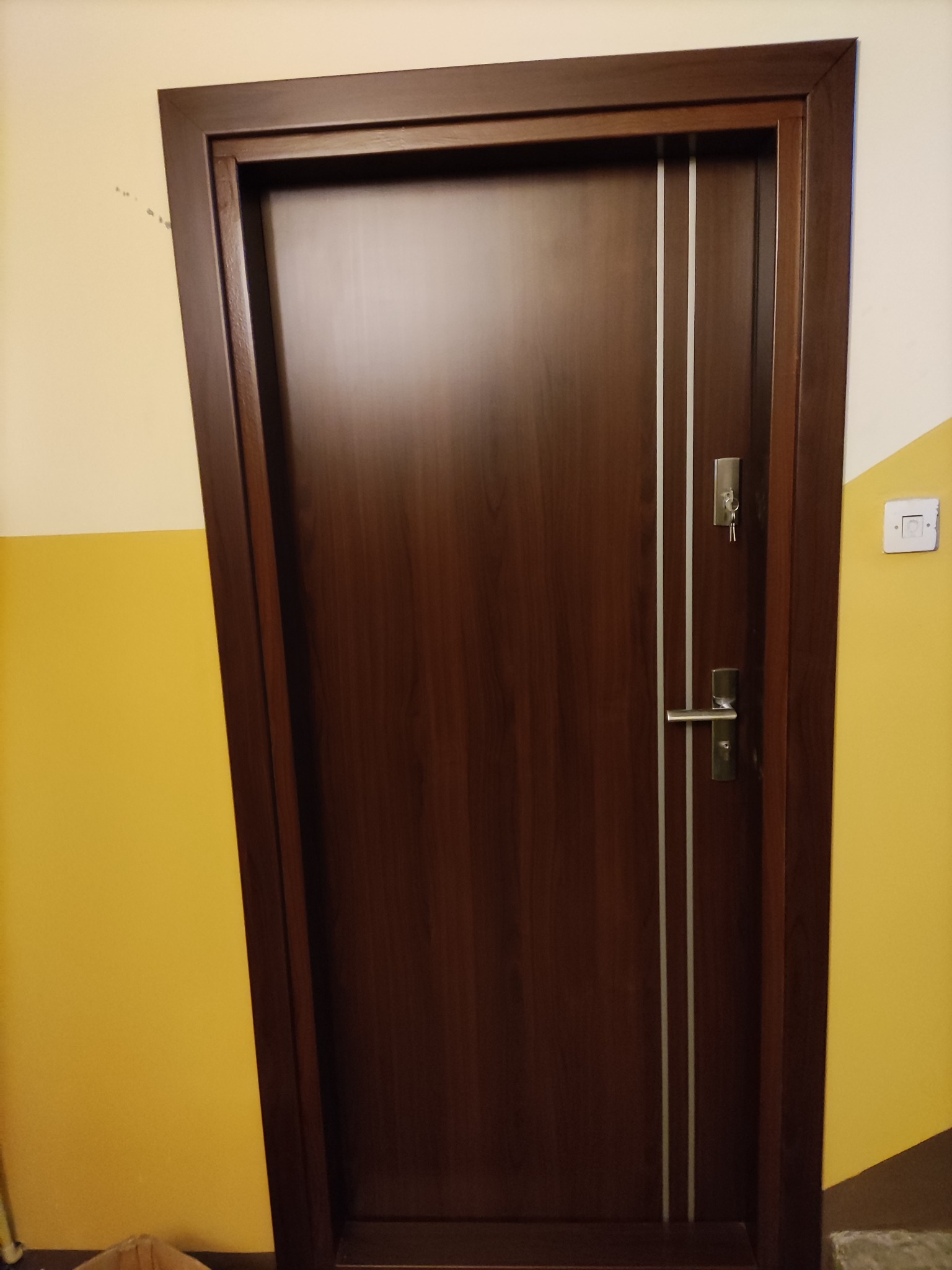 Ciemnobrązowe drzwi z dwoma pionowymi, srebrnymi paskami, zamontowane w ościeżnicy. W zamku włożone klucze. Ściana pomalowana na żółto i biało. Widoczny włącznik światła.