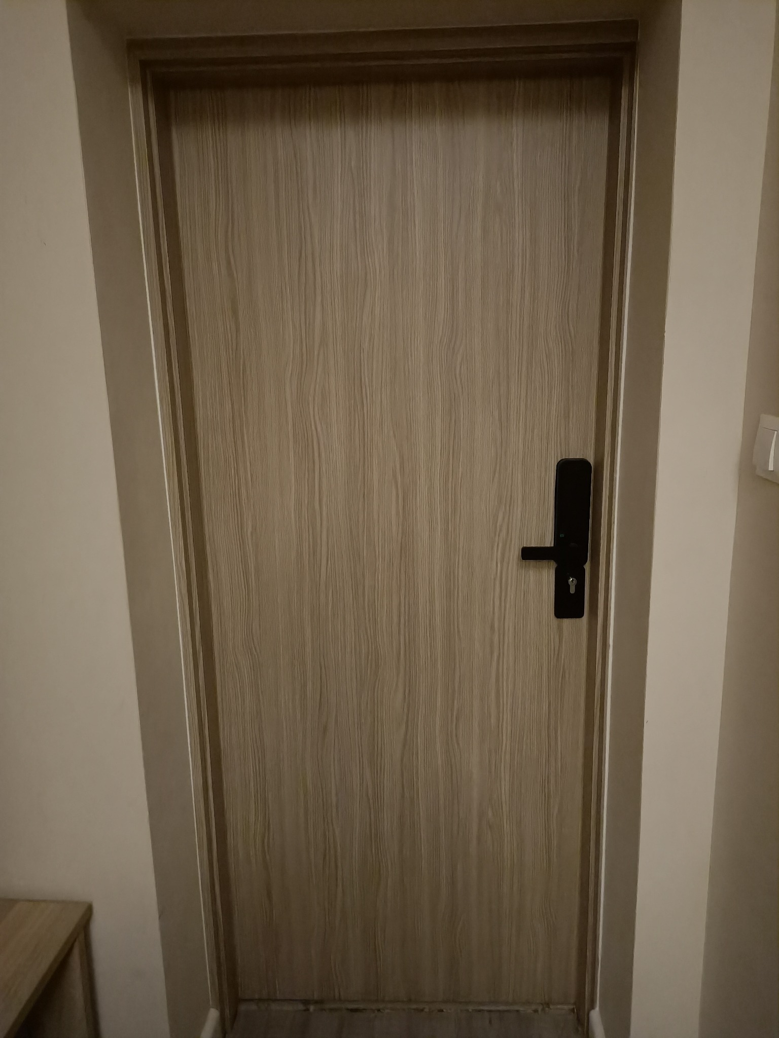 Zamknięte, drewniane drzwi wewnętrzne z czarną, elektroniczną klamką, widoczne w korytarzu o kremowych ścianach.