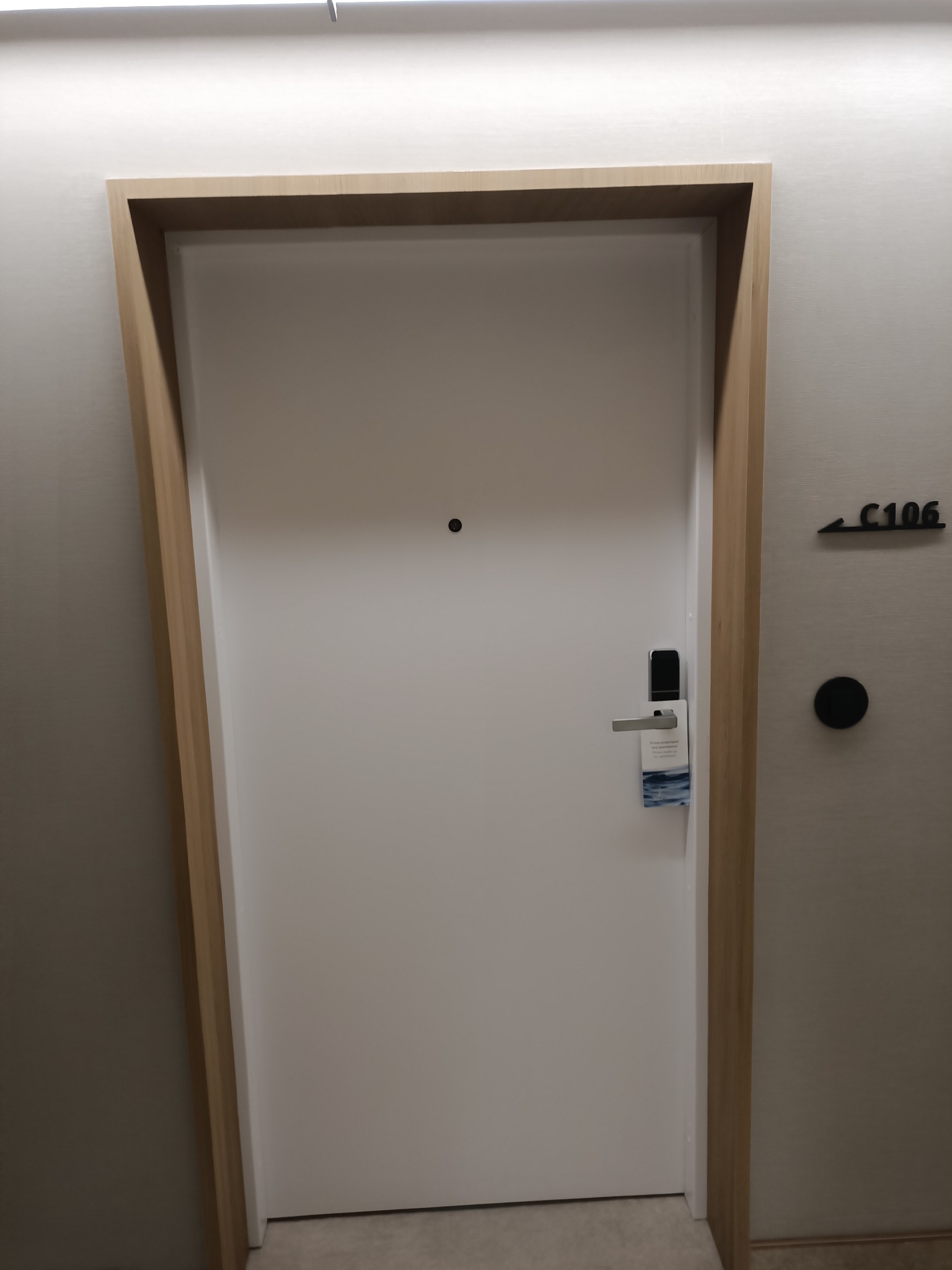 Białe drzwi hotelowe z drewnianą ościeżnicą, elektroniczny zamek na kartę i zawieszka z prośbą o nie przeszkadzanie, widoczny numer pokoju C106.