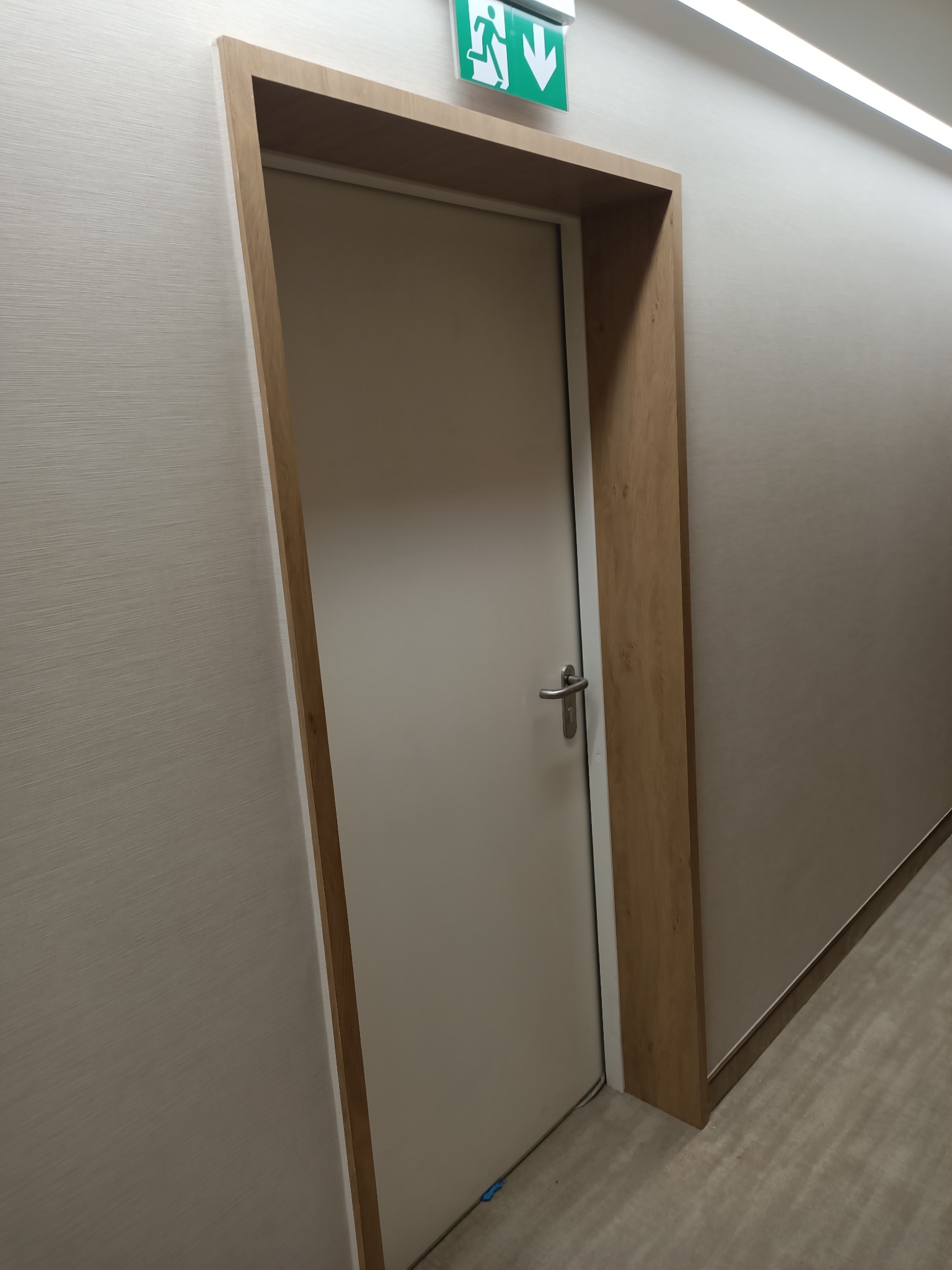 Zamknięte, beżowe drzwi z drewnianą ościeżnicą w korytarzu, nad nimi zielony znak ewakuacyjny.
