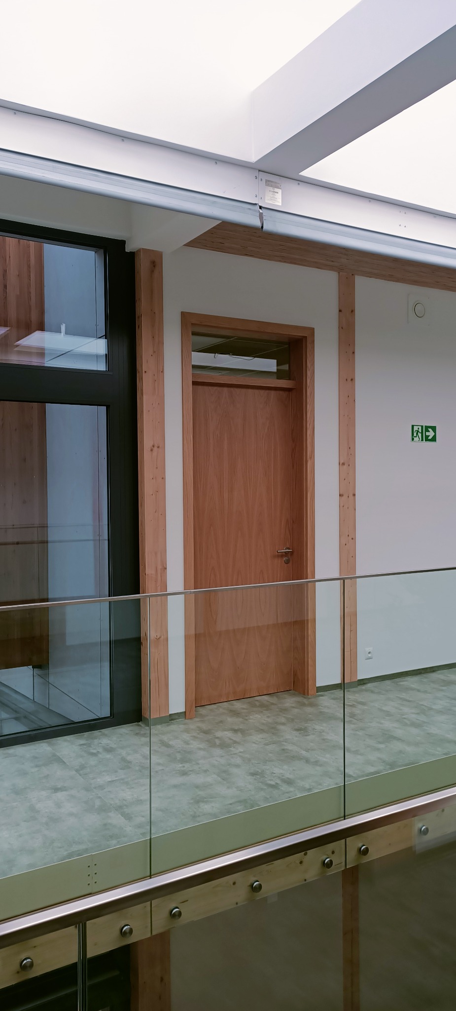 Wnętrze budynku z drewnianą ramą okienną i drzwiami z jasnego drewna, widoczne przez szklaną balustradę. Na ścianie znak ewakuacyjny.