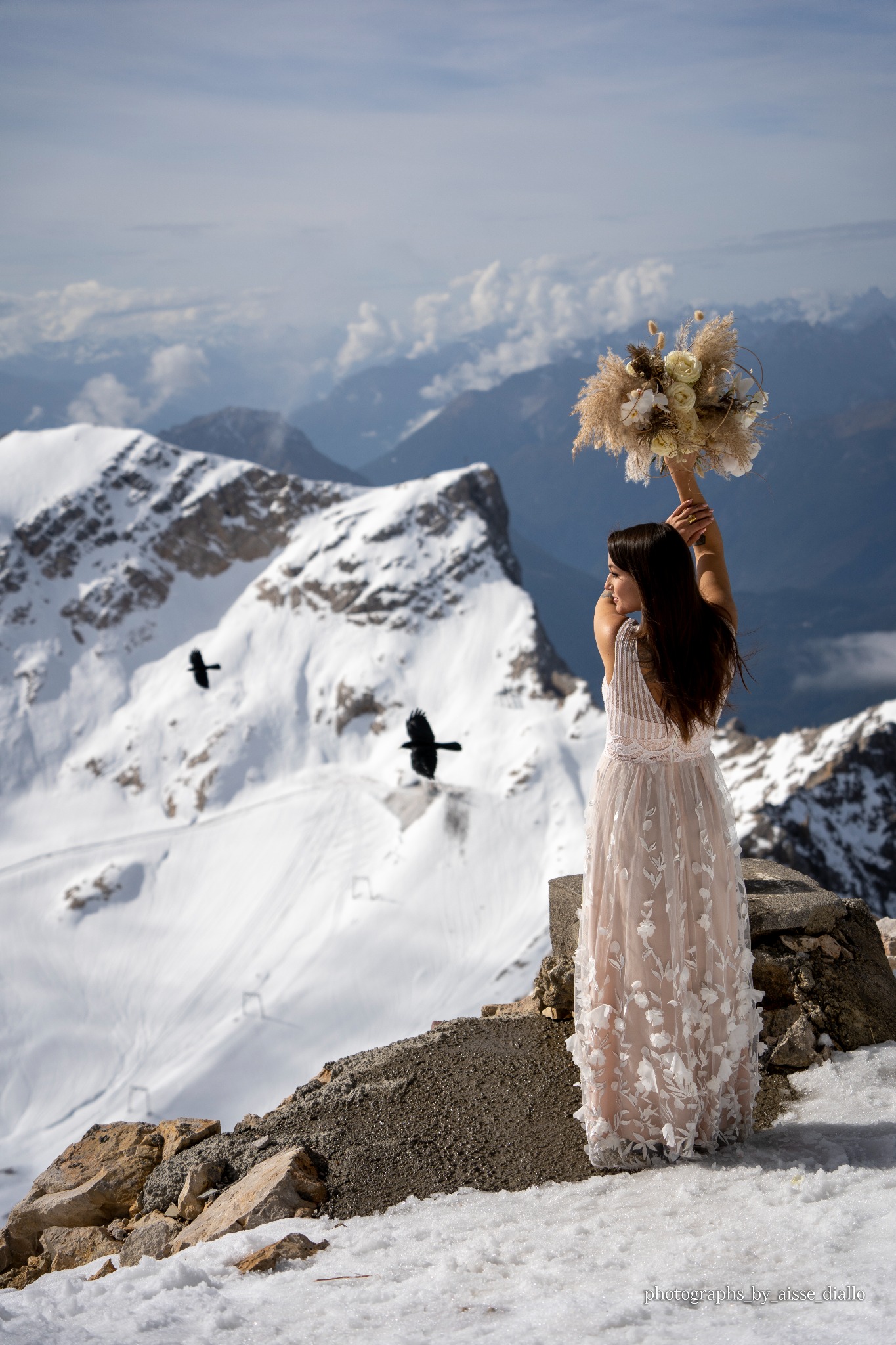 Panna młoda w zwiewnej sukni ślubnej, stojąca na szczycie góry z bukietem wzniesionym do góry; w tle ośnieżone szczyty i dwa lecące kruki.