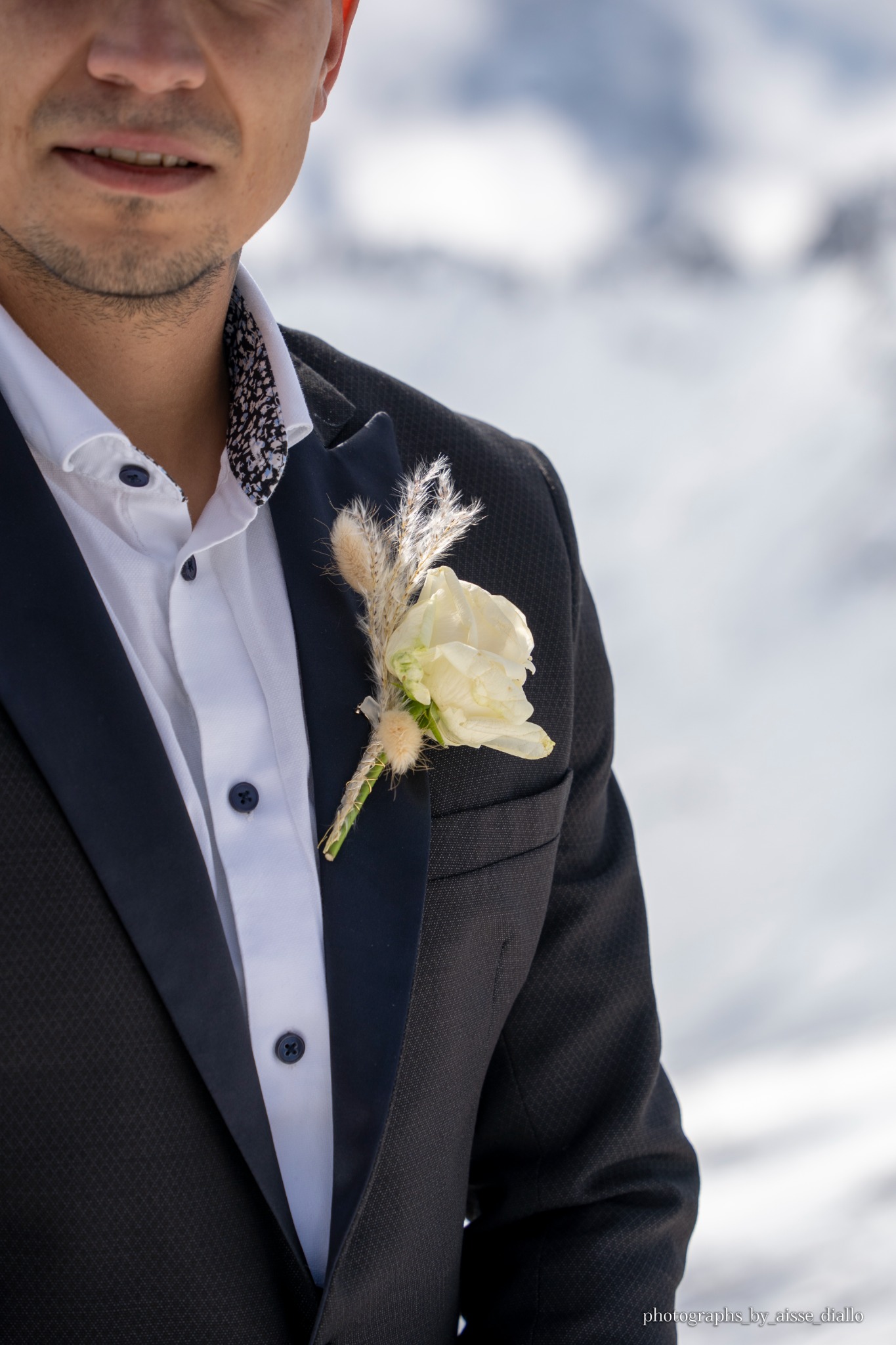 Mężczyzna w garniturze z butonierką z białej róży i traw na tle zimowego krajobrazu.