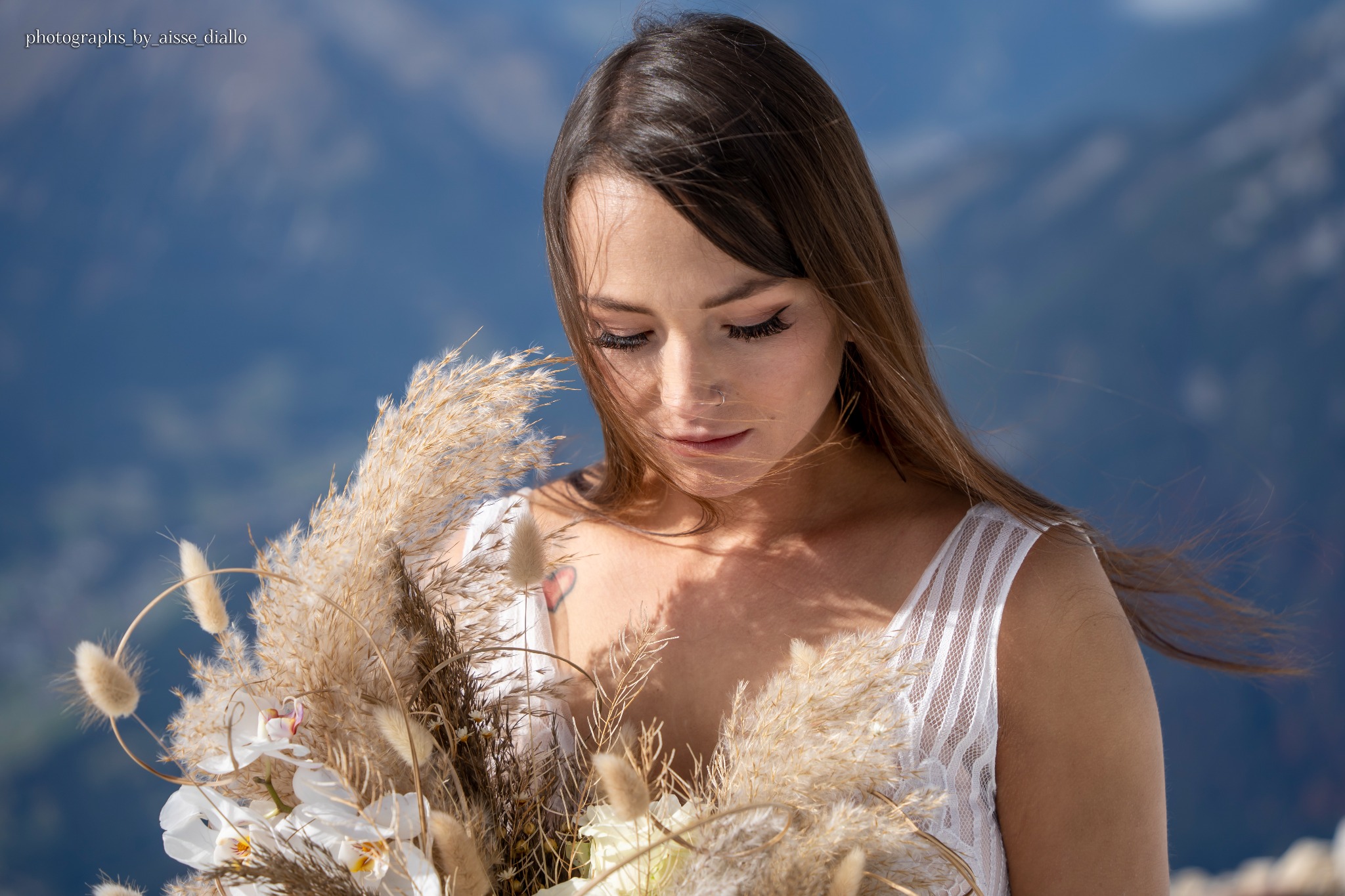 Portret panny młodej z bukietem z traw i białych kwiatów na tle gór, z widocznym tatuażem w kształcie serca na dekolcie.