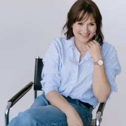 Uśmiechnięta kobieta z brązowymi włosami siedzi na czarnym krześle w niebieskiej koszuli w paski i dżinsach, spoglądając w obiektyw.