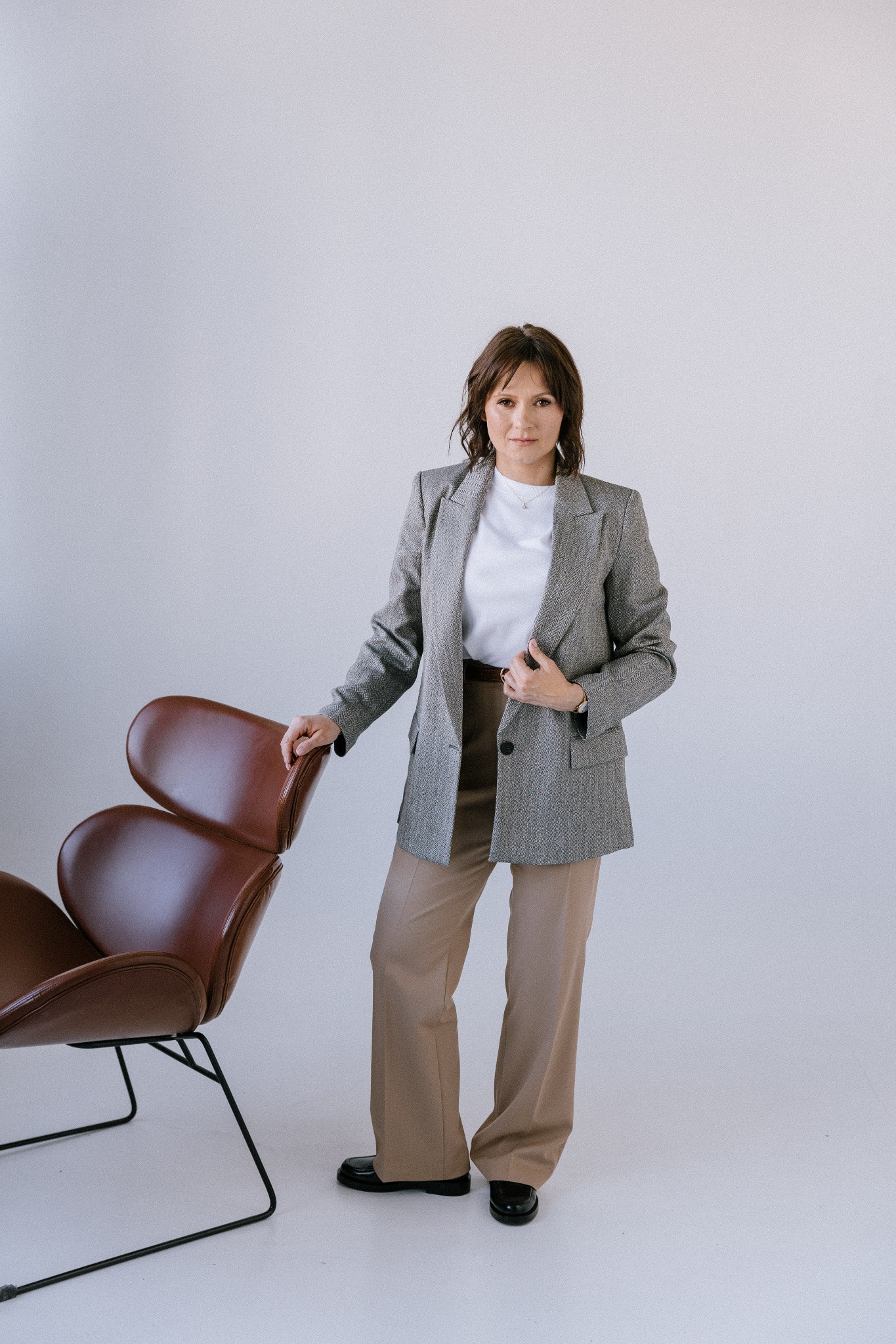 Kobieta w szarym tweedowym żakiecie i beżowych spodniach pozuje obok brązowego fotela o organicznym kształcie na białym tle.
