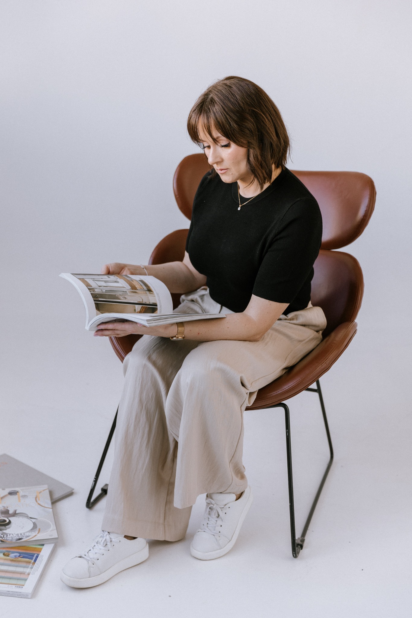 Kobieta siedzi na skórzanym krześle i przegląda magazyn z aranżacjami wnętrz; na podłodze leżą dodatkowe egzemplarze magazynu.