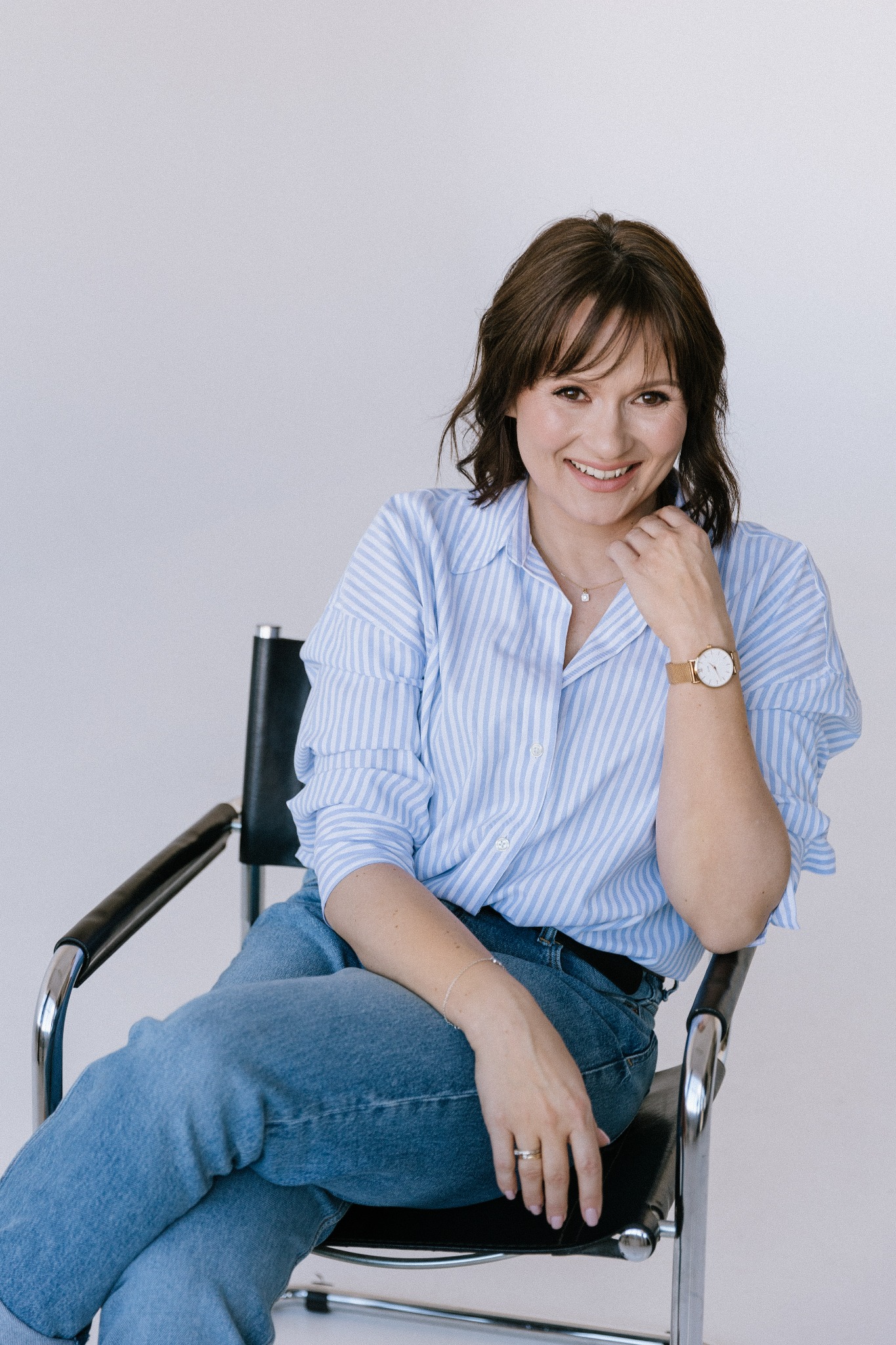 Uśmiechnięta kobieta z brązowymi włosami siedzi na czarnym krześle w niebieskiej koszuli w paski i dżinsach, spoglądając w obiektyw.