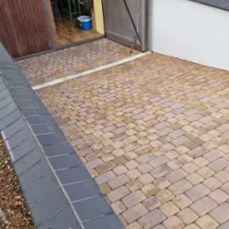 Ułożona kostka brukowa w odcieniach beżu i brązu przed otwartym garażem z drewnianymi drzwiami; widoczny fragment murku z szarymi płytkami.