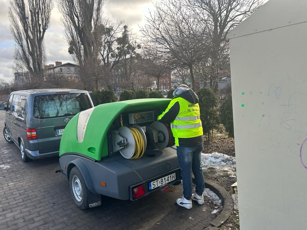 Pracownik w kamizelce odblaskowej obsługuje przyczepę z urządzeniem wysokociśnieniowym DIBO na kostce brukowej, obok zaparkowany szary Volkswagen Transporter.