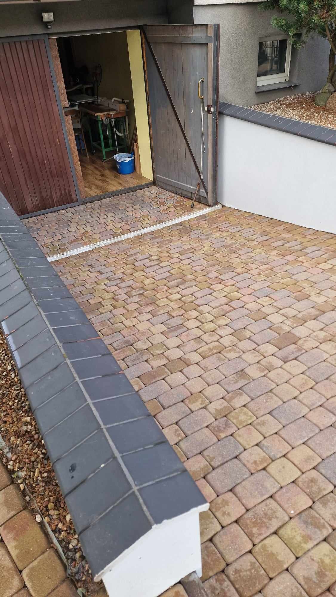 Ułożona kostka brukowa w odcieniach beżu i brązu przed otwartym garażem z drewnianymi drzwiami; widoczny fragment murku z szarymi płytkami.