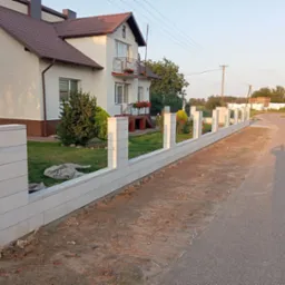 Nowoczesne ogrodzenie z białych, modułowych bloczków betonowych, oddzielające posesję z domem jednorodzinnym od drogi asfaltowej, w tle widoczny cień fotografa.
