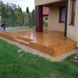 Nowo wybudowany drewniany taras z dwoma stopniami, przylegający do domu z ceglanym filarem, w trakcie czyszczenia przez pracownika, z widocznymi śladami piasku i zabrudzeń po budowie.