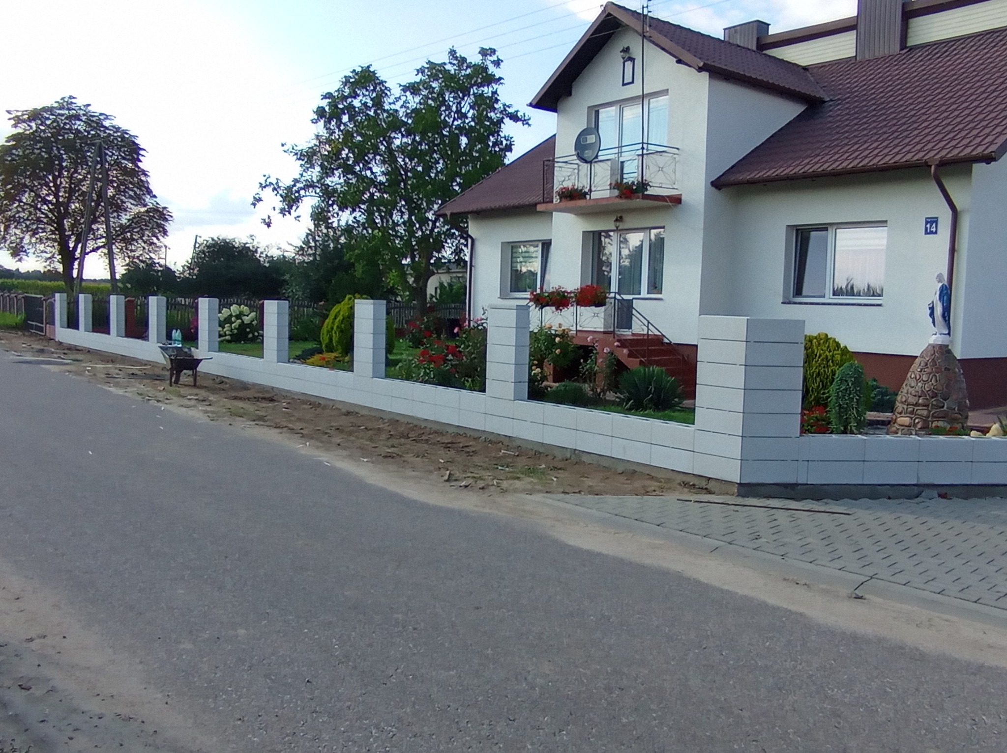 Nowoczesne ogrodzenie z szarej kostki betonowej z białymi przęsłami, z widocznym fragmentem ulicy i domu z ogrodem, w tle drzewa i niebo.