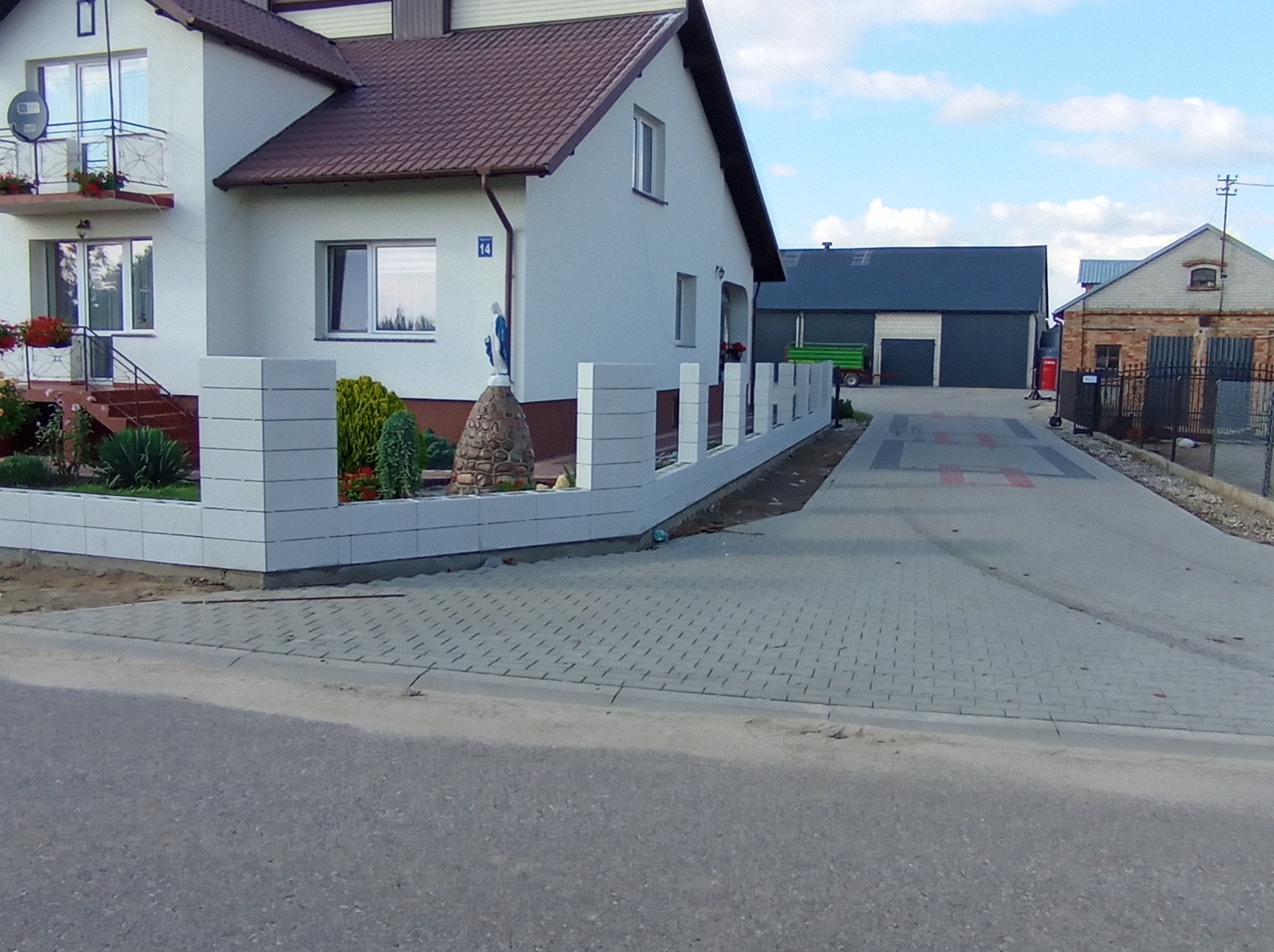 Nowoczesne ogrodzenie z białych, betonowych bloczków otaczające posesję z kostką brukową na podjeździe. Na trawniku niewielka figurka Matki Boskiej.