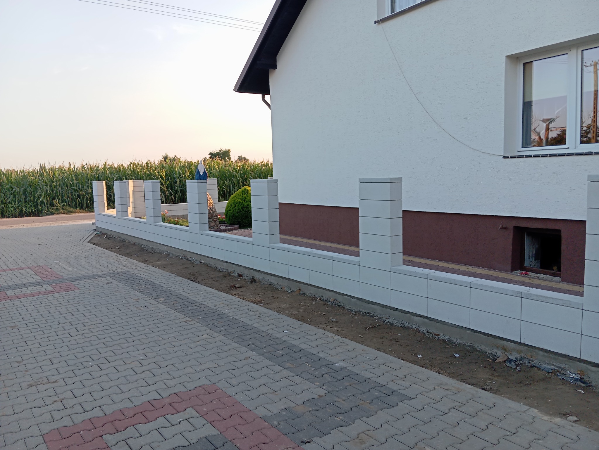 Nowoczesne ogrodzenie z białych, modułowych bloczków betonowych z pustakami, oddzielające posesję od chodnika z kostki brukowej. W tle pole kukurydzy i fragment domu z oknem.