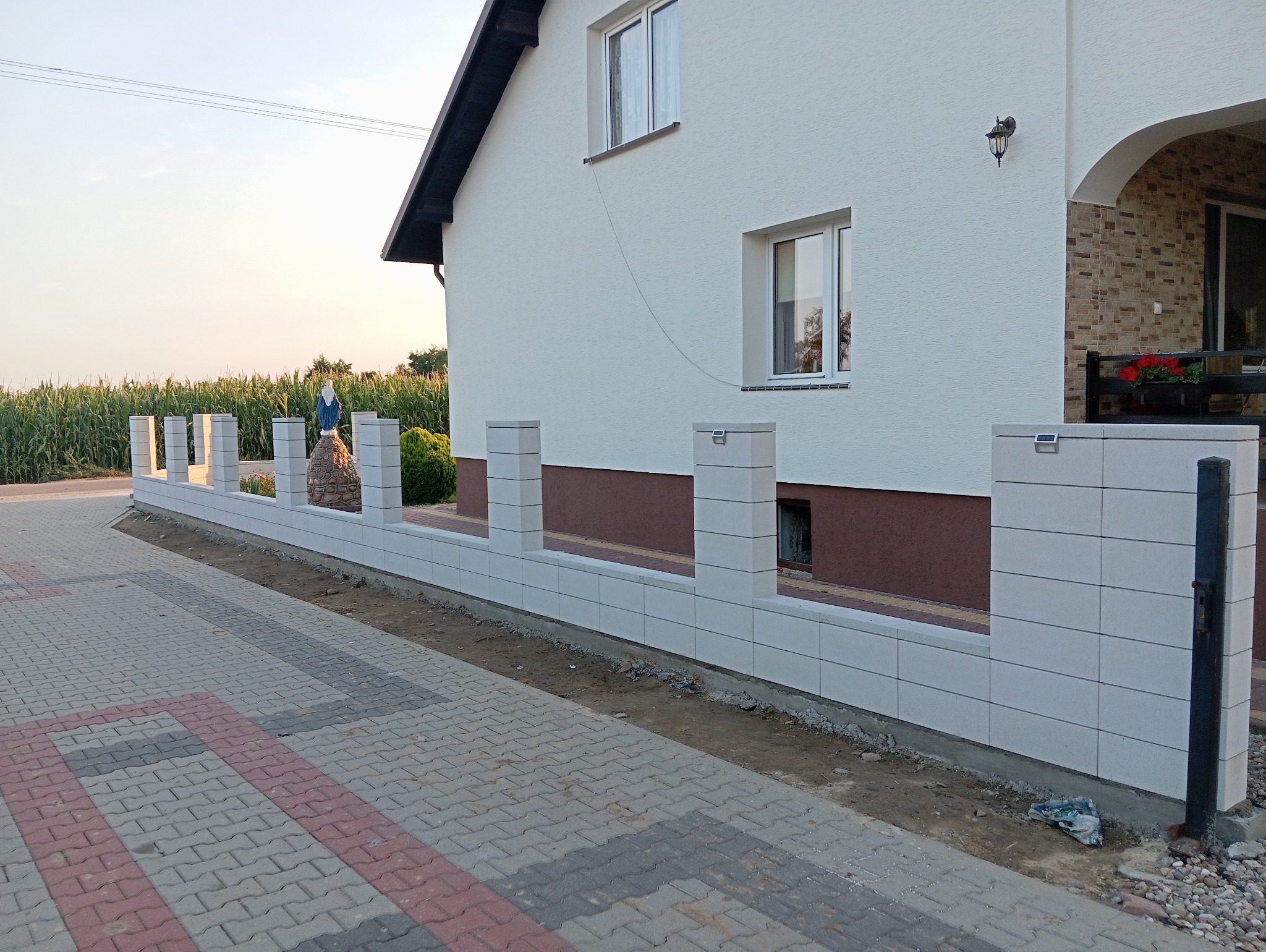Nowoczesne ogrodzenie modułowe z białych bloczków betonowych, zakończone słupkami z oświetleniem solarnym, otaczające posesję z widoczną figurką Matki Boskiej w tle pola kukurydzy.