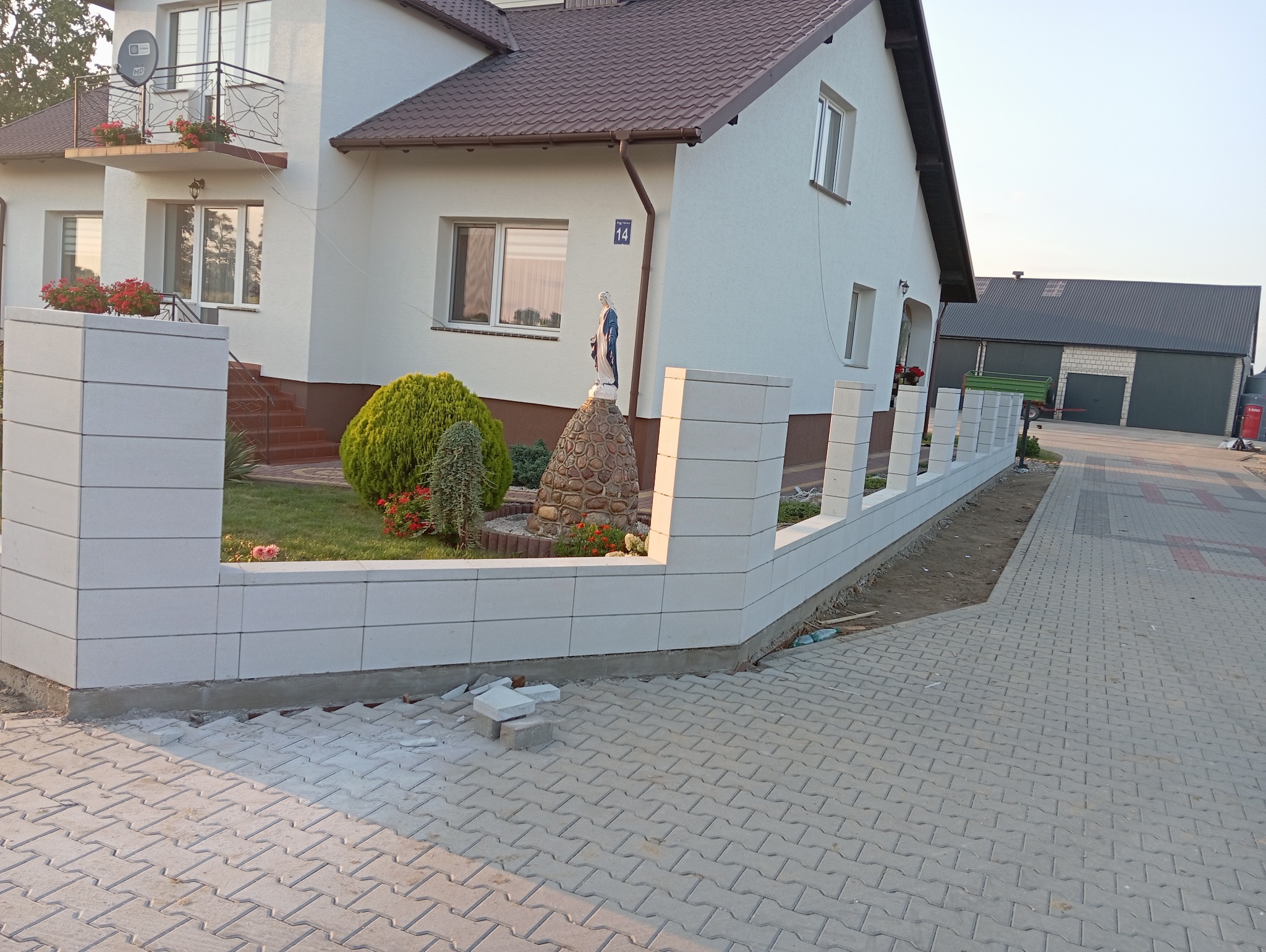 Nowoczesne ogrodzenie z białych, modułowych elementów betonowych, otaczające posesję z kostką brukową, widok na dom z balkonem i figurką Matki Boskiej w ogrodzie.