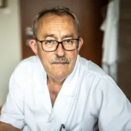 Portret starszego mężczyzny w okularach i białym fartuchu medycznym, z wąsami, siedzącego w gabinecie.