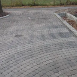 Szara kostka brukowa ułożona na podjeździe, z obrzeżami z kostki granitowej. Widoczna zieleń trawnika w tle. Kąt ujęcia z góry, podkreślający wzór ułożenia kostki.