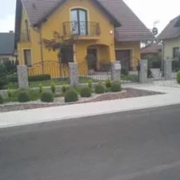 Żółty dom jednorodzinny z brązowym dachem, ozdobny metalowy płot z kamiennymi słupkami i zadbany ogród z kulistymi krzewami oraz chodnikiem z kostki.