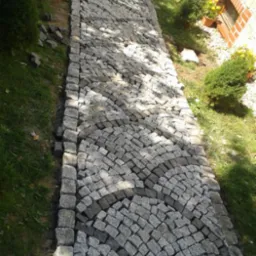 Ścieżka z kostki granitowej ułożona w łukowaty wz&oacute;r, obramowana regularnymi kostkami. Trawa i małe drzewka ozdabiają otoczenie.