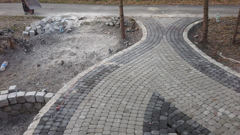 Ścieżka z kostki granitowej w odcieniach szarości, układana łukowo, z widocznymi zapasami kostki i fragmentem trawnika. Etap prac w toku.
