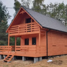 Nowoczesny, dwukondygnacyjny budynek z drewna, z balkonem i ciemnym dachem, posadowiony na betonowych bloczkach, na tle drzew.