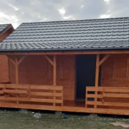 Nowo wybudowany, drewniany domek letniskowy z ciemnoszarą dachówką, widoczny z przodu z częściowo zadaszonym tarasem z drewnianą balustradą, umieszczony na betonowych bloczkach.