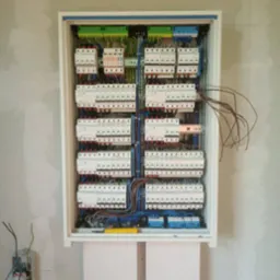 Rozdzielnica elektryczna w trakcie montażu w surowym wnętrzu, widoczne liczne zabezpieczenia, przewody w różnych kolorach i niepodłączone kable, na ścianie widoczny puszkowy wyłącznik.