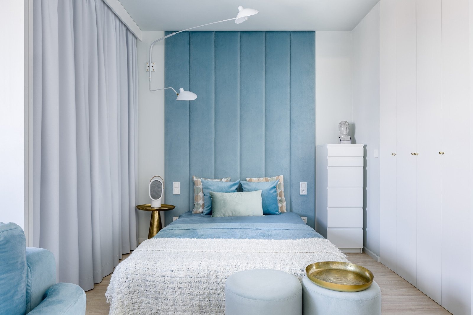 Nowoczesna sypialnia w odcieniach błękitu i bieli, z tapicerowanym panelem ściennym, białymi szafami i złotymi akcentami, takimi jak stolik nocny i taca dekoracyjna.