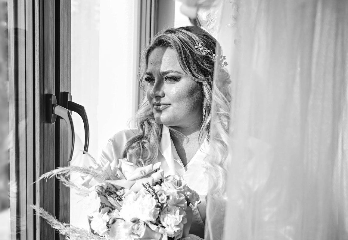 Czarno-białe zdjęcie panny młodej w satynowym szlafroku, z bukietem kwiatów, stojącej przy oknie z drewnianą ramą i metalową klamką, z delikatną ozdobą we włosach i długimi kolczykami...