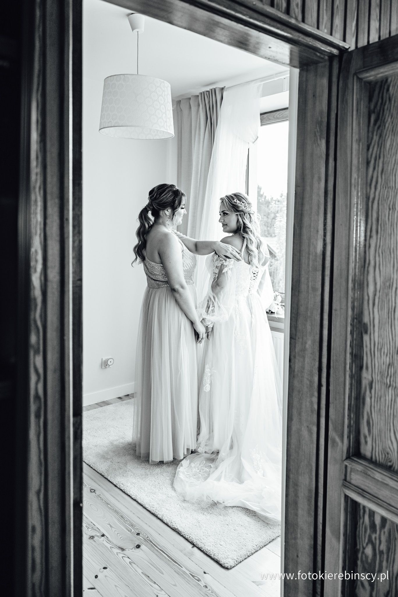 Czarno-białe zdjęcie panny młodej w koronkowej sukni ślubnej, pomagającej jej druhny w dopinaniu sukni w pokoju z oknem i lampą.
