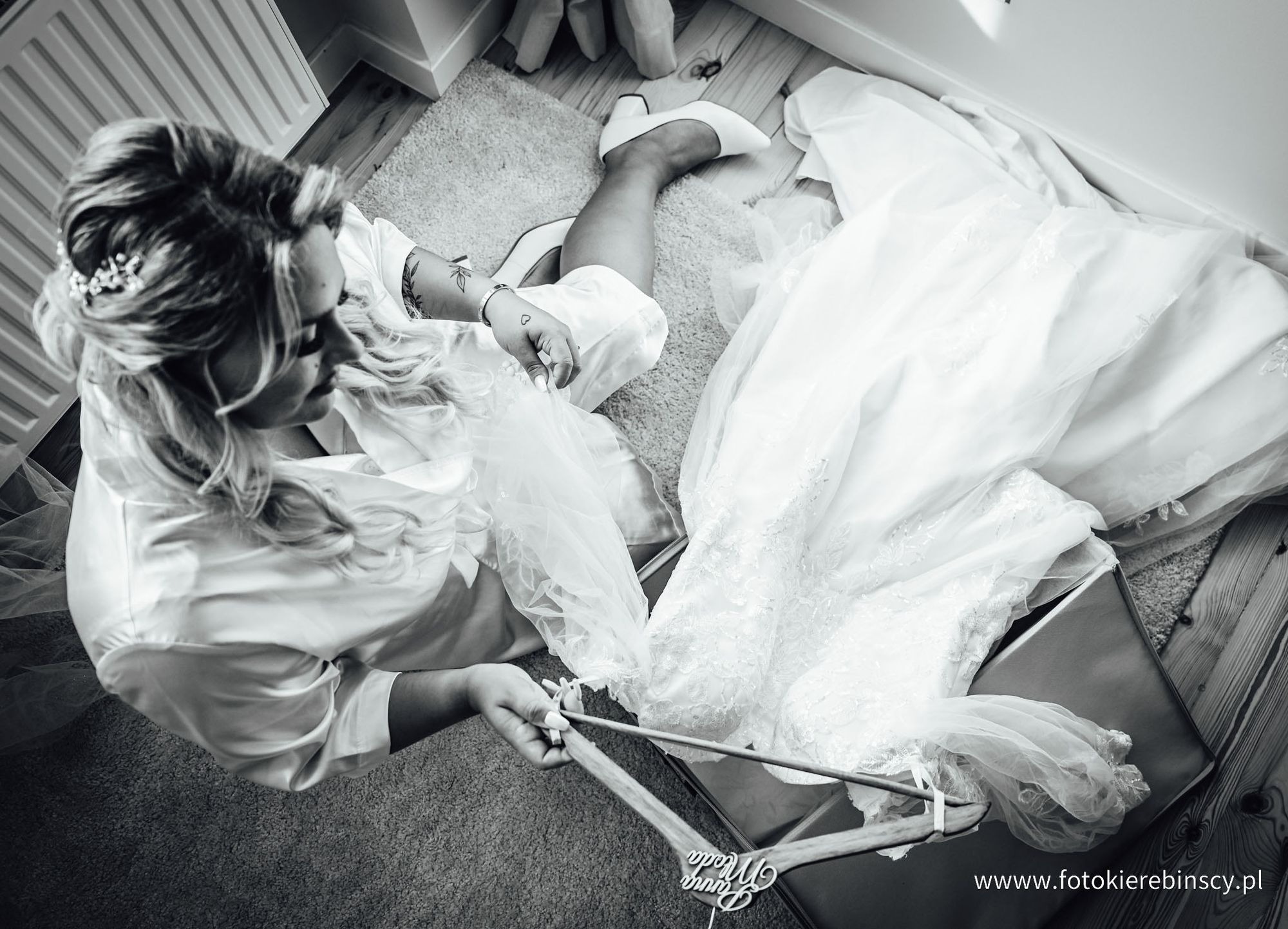 Czarno-białe zdjęcie z góry przedstawia pannę młodą w szlafroku, przygotowującą suknię ślubną z koronkowymi detalami, leżącą na pudełku i wieszaku z napisem 'Panna Młoda'. Widoczne białe szpilki...